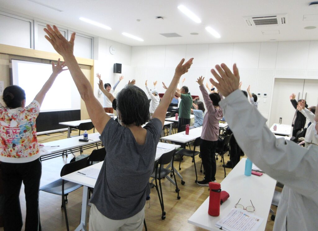市原市「笑って健康講座」で、参加者が両手を大きく伸ばしながら健康体操を楽しんでいる様子。