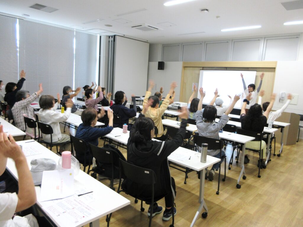 市原市「笑って健康講座」で、参加者が座ったまま手を動かしながら笑顔で健康体操を楽しんでいる様子。